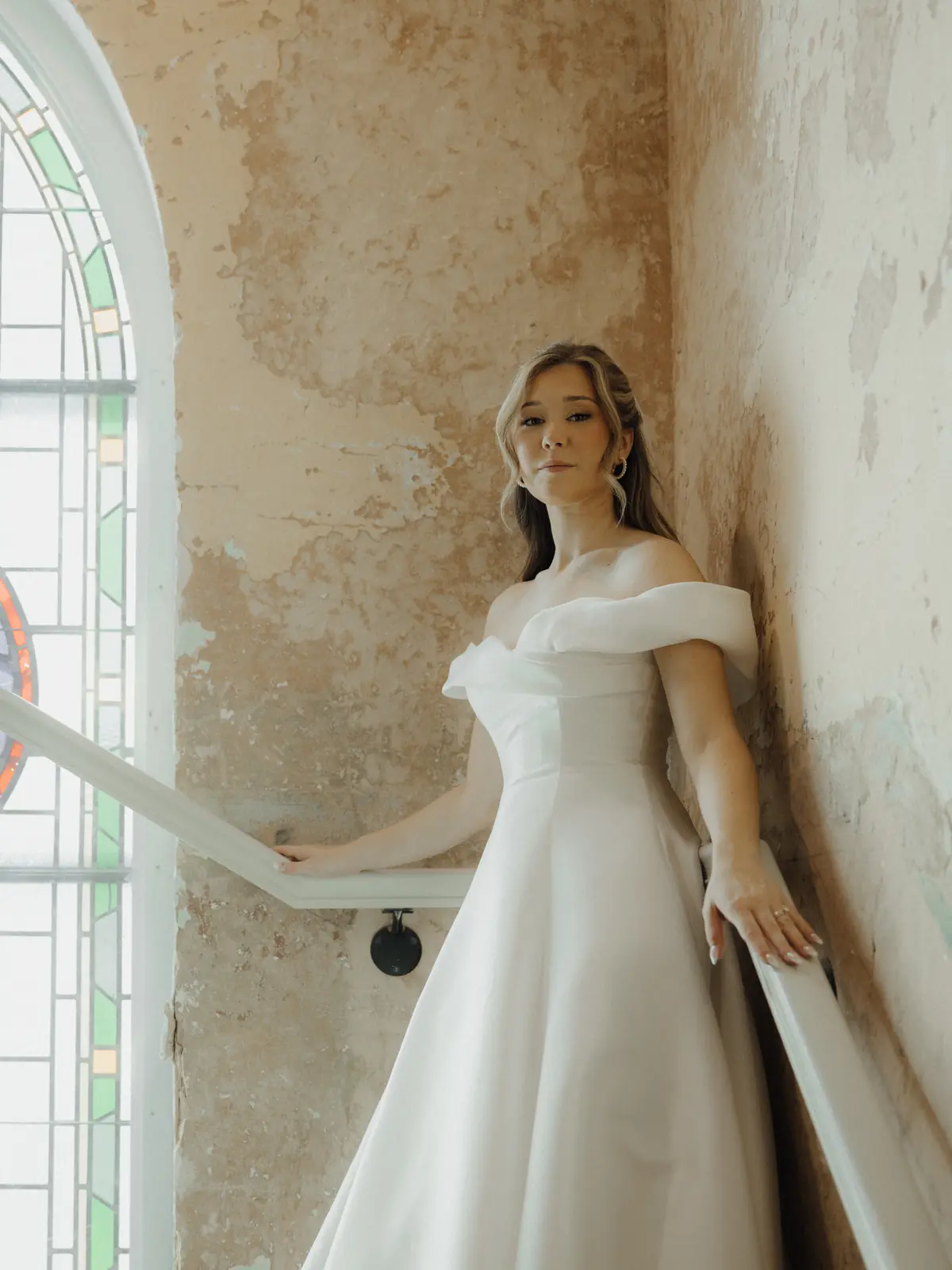 Bride in an off-shoulder gown leaning in a sunlit old stairwell